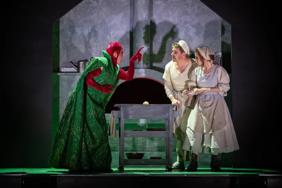 The Scoop Stage scene: a witch in a green robe and red hood points at two costumed villagers by a wooden table and cauldron in a gray kitchen set.